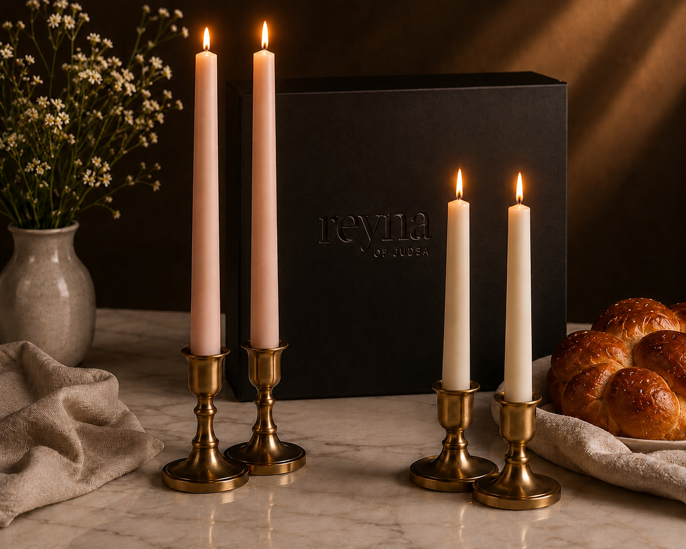 Four lit taper candles in brass candlesticks arranged on a marble surface, with a black Revia gift box behind them and decorative bread and flowers nearby.