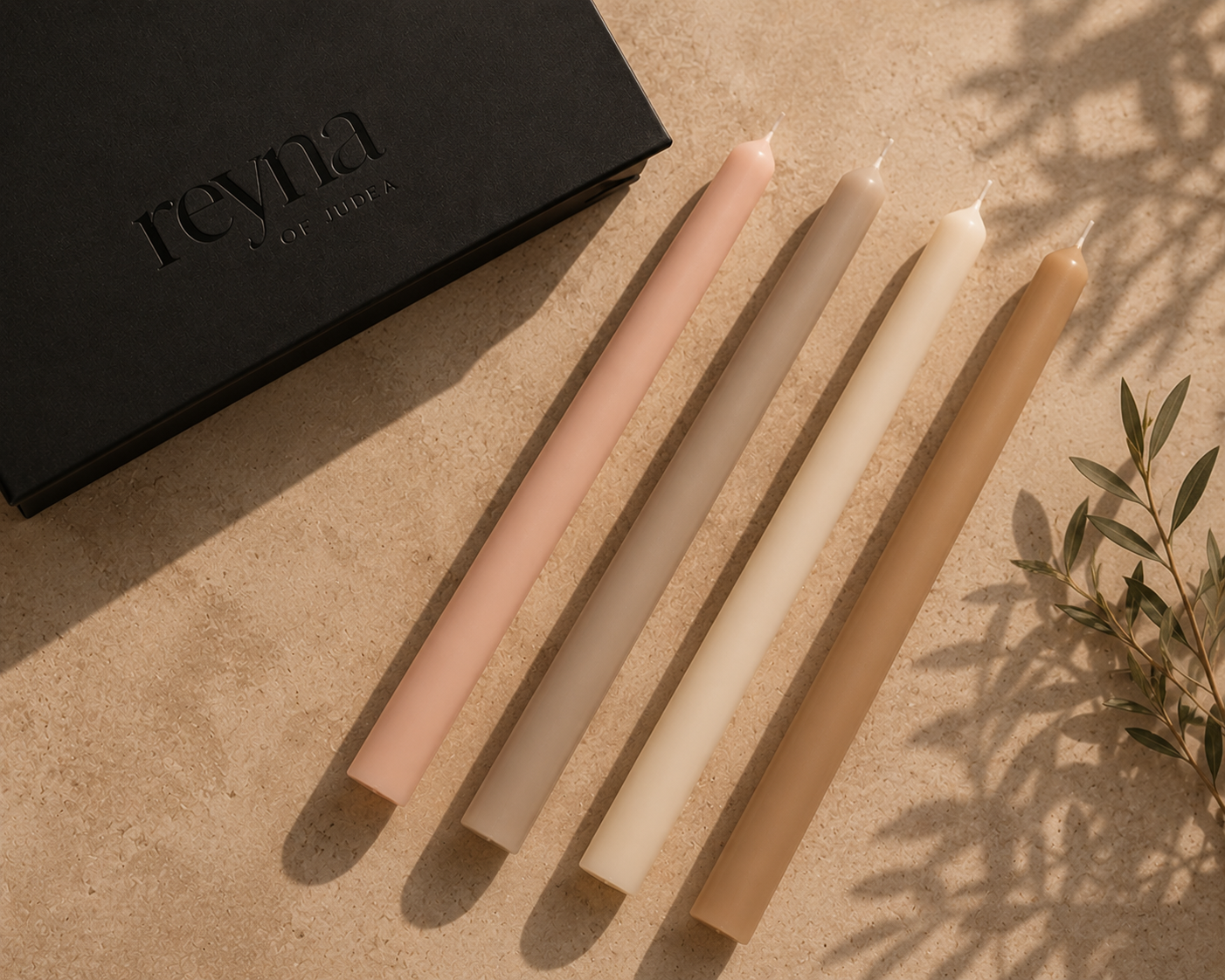 Four taper candles in soft neutral shades of blush pink, taupe, cream, and tan, arranged diagonally next to a black Revna gift box on a textured beige surface with leaf shadows.